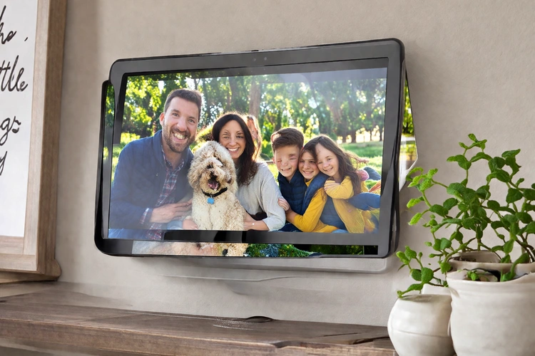 A tablet mounted on a wall or propped on a shelf displaying a rotating slideshow of family photos in a living room.