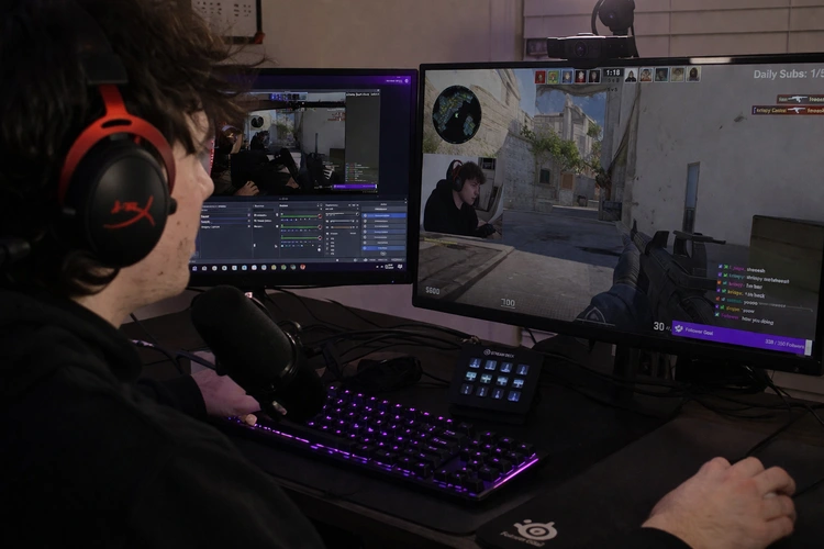 A gamer live streaming on Twitch with a webcam showing their face in the corner overlay, OBS Studio visible on a second monitor. Gaming setup with RGB keyboard and headset.