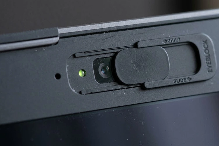 A thin webcam privacy shutter cover sliding over a laptop's built-in camera lens. Close-up macro shot showing the physical slide mechanism against a dark laptop bezel.