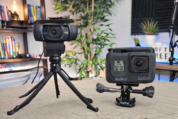 A USB webcam on a desk tripod placed next to a compact action camera on a small mount, both aimed at the same angle in a content creator home studio setup.